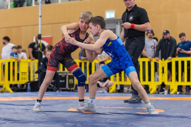 Two young wrestlers in red and blue singlets lock grips on the mat, focused and determined during a youth bout.