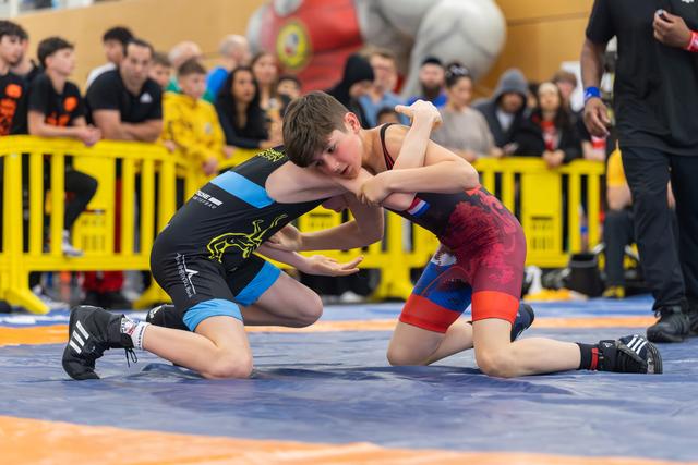 Two young wrestlers locked in a grappling duel on a blue mat, both focused and determined, crowd watching closely behind barriers.