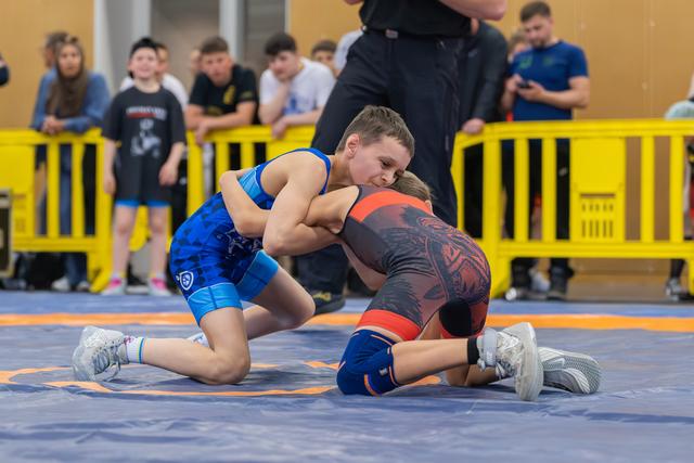 Two youth wrestlers locked in a fierce grapple on a blue mat, blue singlet athlete gaining top control over opponent.