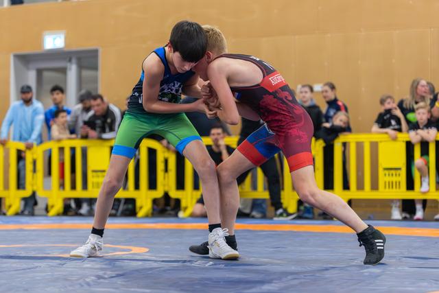 Two young boys in wrestling singlets grip each other intensely on a blue mat, surrounded by spectators behind yellow barriers.