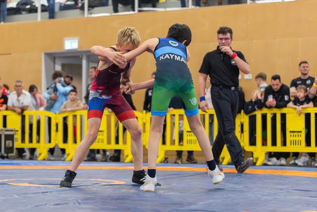 Two young boys in singlets grip each other intensely on a blue mat, referee watching closely behind yellow barriers.