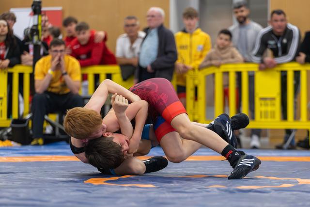 Two young wrestlers locked in a ground hold on the mat, crowd of coaches and spectators watching intently behind yellow barriers.