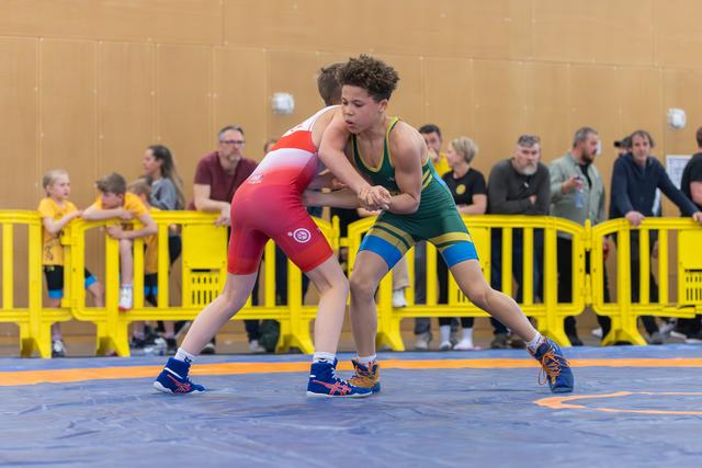 Two young wrestlers lock grips on the mat, the green-suited athlete driving forward with focused determination against his red-suited opponent.
