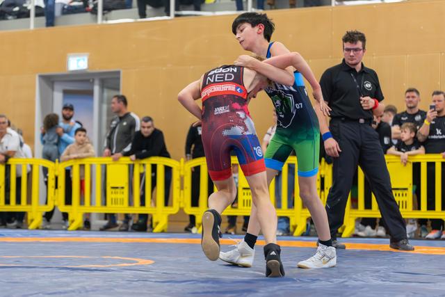 Two young wrestlers grapple intensely on the mat, one in a NED singlet, referee watching nearby.