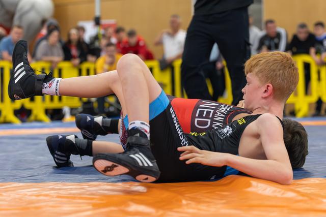 Two young boys wrestle on an orange mat, one pinning the other in a controlled hold during an indoor event.