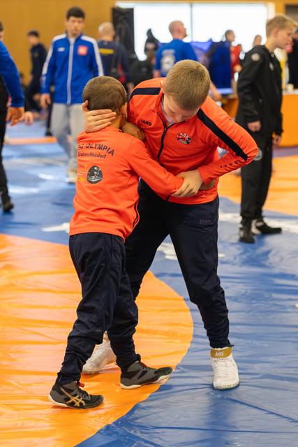 Two boys in orange AC Olympia jackets clinch during a wrestling bout on orange and blue mats, focused and intense.