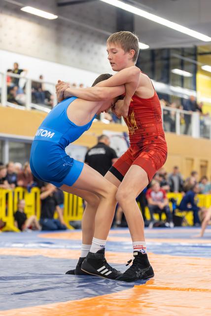 Two youth wrestlers locked in an intense clinch, red vs blue singlets, competing on an orange mat before a crowd.