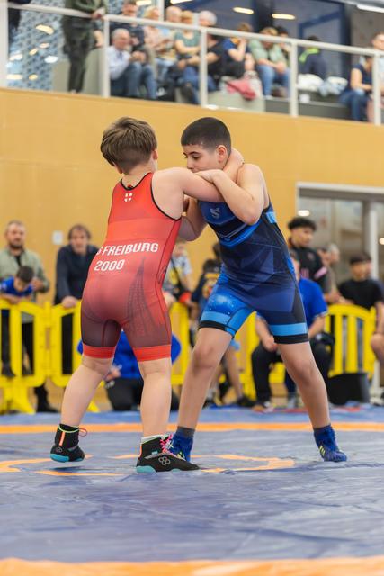 Two young wrestlers in red and blue singlets engage in a grappling match on a blue mat, with spectators watching.