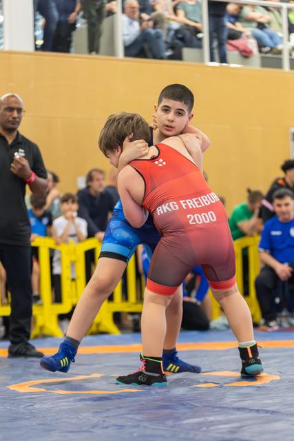 Two youth wrestlers in a tight clinch on the mat; red singlet reads RKG Freiburg 2000, both focused and determined.