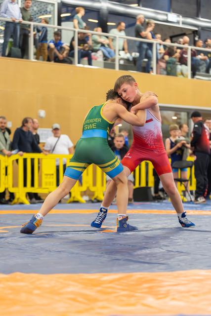 Swedish wrestler Verdier in green grapples intensely with a red-suited opponent on a blue mat before a crowded arena.