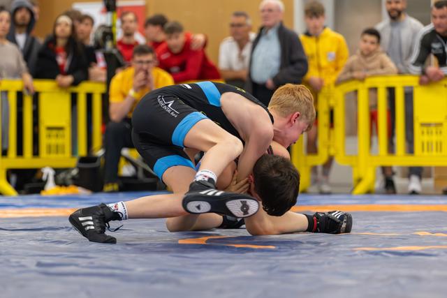 Young red-haired wrestler dominates opponent, driving him to the mat with intensity before a watching crowd.