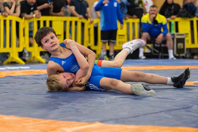 Young boy in blue Adidas singlet pins opponent on mat, looking up with focus during youth wrestling bout.
