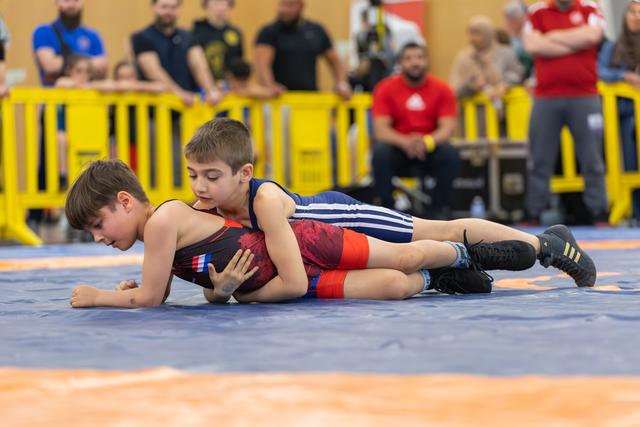 Young boy in blue singlet pins opponent to the mat, focused expression, amid a crowd of spectators at an indoor event.