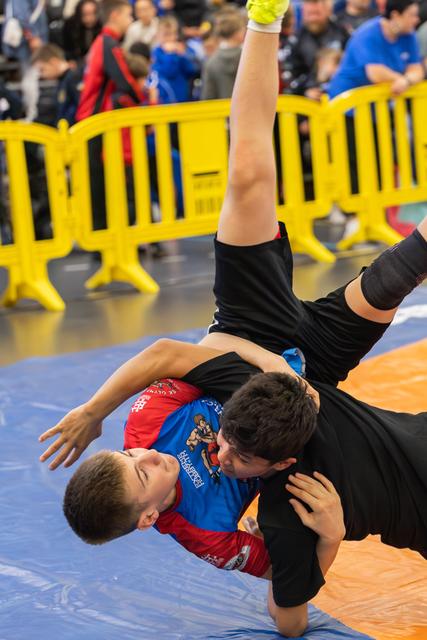 Two young wrestlers grapple intensely on a blue mat, one flipping the other mid-air during a close match.