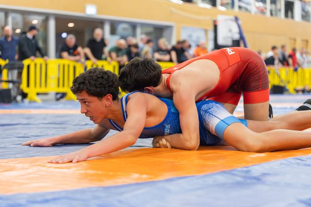 Young wrestler in blue singlet, pinned down by opponent in red, holds composure with focused determination on orange mat.