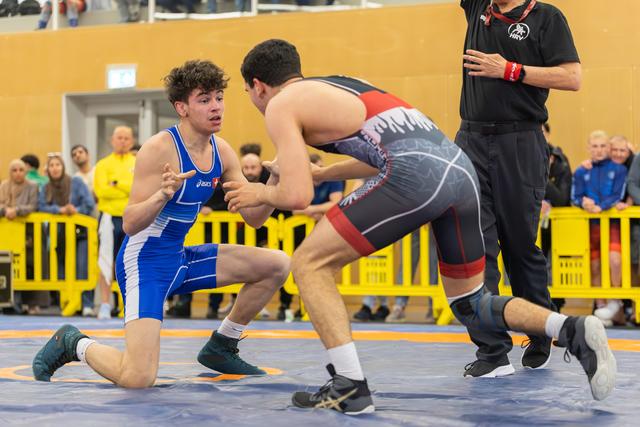 Young wrestler in blue Asics singlet crouches defensively on one knee, eyes locked on opponent in black-red singlet.