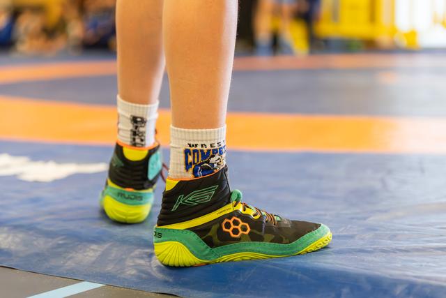 Close-up of a young wrestler's feet in vibrant Rudis shoes and wrestling socks, standing on a blue competition mat.
