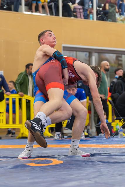 Young wrestler in red singlet lifts opponent off the mat, muscles straining, crowd watching from the stands behind.