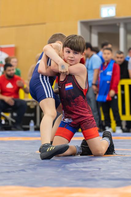 Young Dutch wrestler in red singlet locks opponent's arms, focused expression, kneeling on blue mat at indoor youth event.