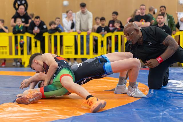 Young wrestler pins opponent on orange mat as a focused coach kneels nearby, spectators watching behind yellow barriers.