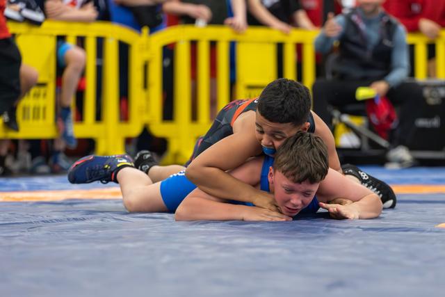 Young boy in blue singlet pinned to the mat, grimacing, as opponent controls him from above on blue wrestling mat.