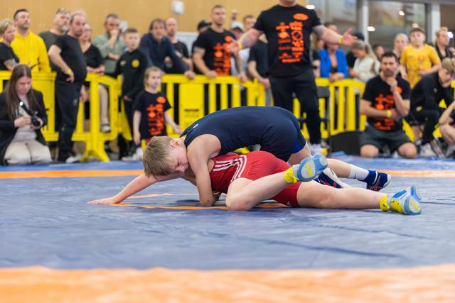 Young wrestler in red singlet being pinned by opponent in navy blue, crowd watching intently in background.