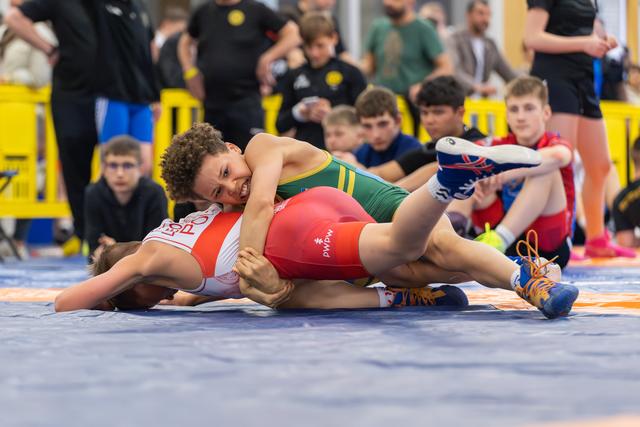 Young wrestler in green singlet pins opponent to the mat, grimacing with effort during a youth wrestling match.