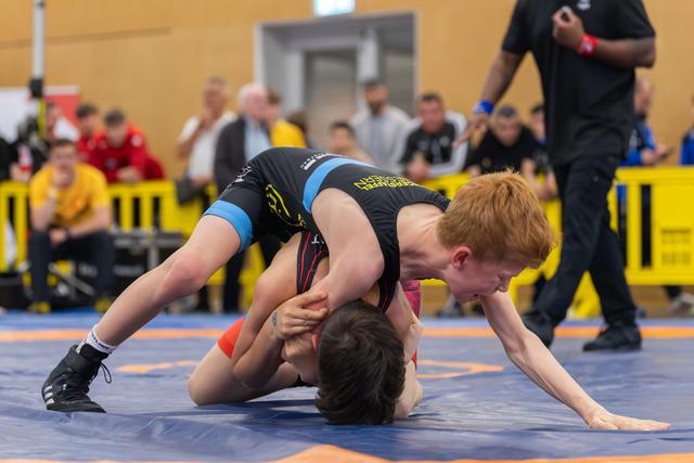 Red-haired boy dominates his opponent on the mat, pressing him down with focused intensity during a youth match.