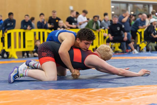 Young wrestler with curly hair controls blonde opponent on blue mat, leaning in with intense focus during indoor match.