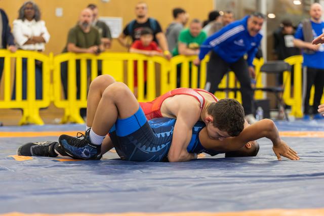 Young wrestler in red singlet pins opponent in blue to the mat during an indoor match, coach watching intently nearby.