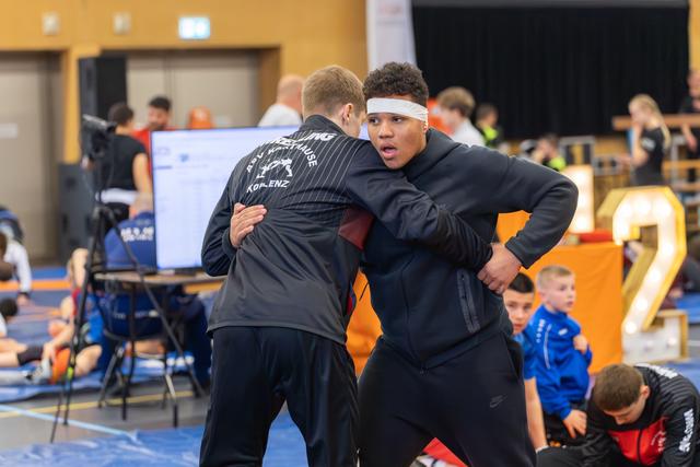Two young wrestlers lock into a clinch grip on the mat, the taller boy in a white headband showing focused intensity.