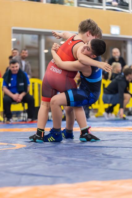 Two young wrestlers locked in a tight clinch on a blue mat, both straining with effort during a competitive bout.