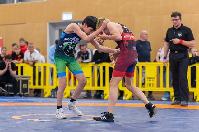 Two young wrestlers lock grips on a blue mat, both leaning in intensely, referee watching closely behind yellow barriers.
