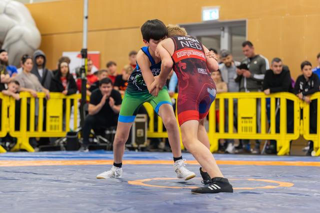 Two young wrestlers locked in a grapple on a blue mat, crowds watching behind yellow barriers at an indoor sports hall.