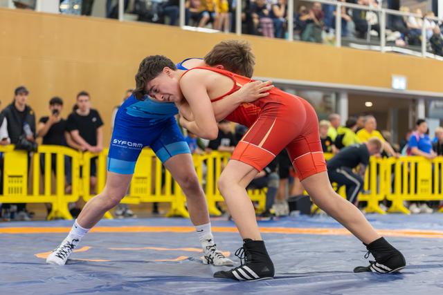 Two young wrestlers lock up on the mat, both focused and determined, competing in blue and red singlets at an indoor event.