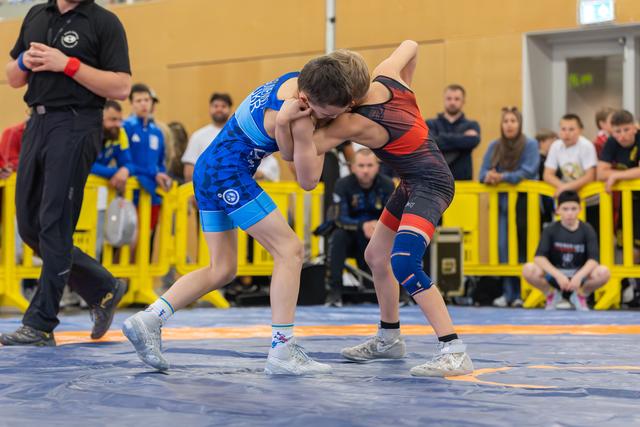 Two young wrestlers locked in a grappling hold, both leaning forward intensely on a blue competition mat with spectators watching.