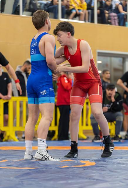 Two young wrestlers engage in a standing clinch, the red-suited boy grips his Swiss opponent with focused intensity on the mat.