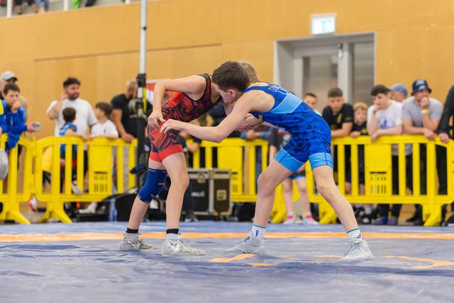 Two young wrestlers in blue and red singlets lock grips on the mat, focused and tense during a youth bout.
