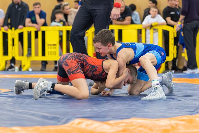 Two young wrestlers compete intensely on a blue mat, one in red dominating over the other in blue during a youth bout.