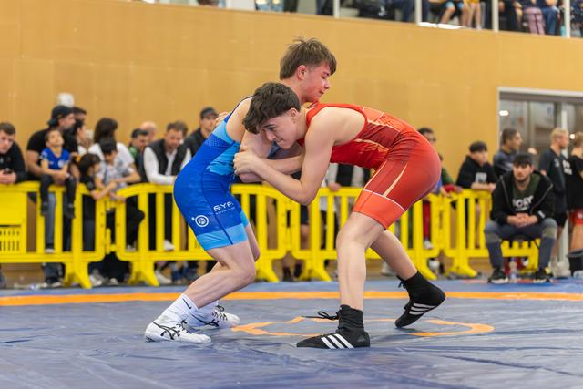 Two young wrestlers locked in a fierce clinch on the mat, spectators watching from behind yellow barriers.