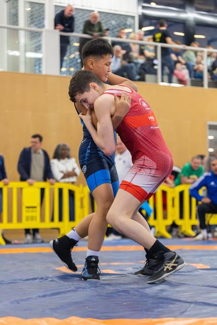 Two young wrestlers locked in a tight clinch on the mat, both straining with intense focus during a youth bout.