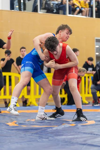 Two young wrestlers grapple fiercely on a blue mat, one in blue lunging forward, the other in red bracing defensively.