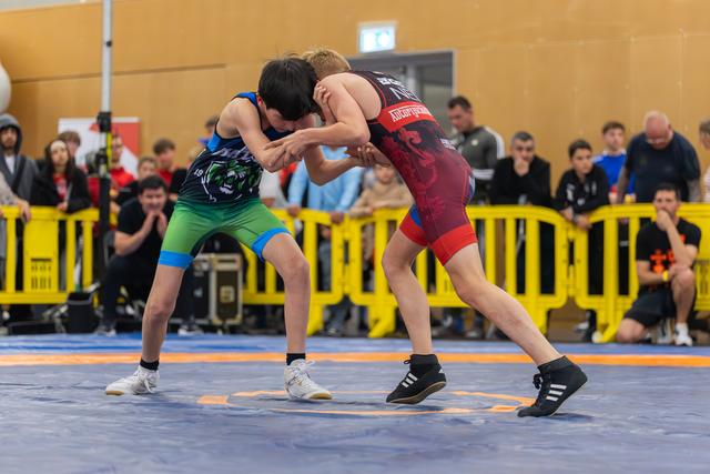 Two young wrestlers lock in a tight clinch on a blue mat, surrounded by spectators behind yellow barriers.
