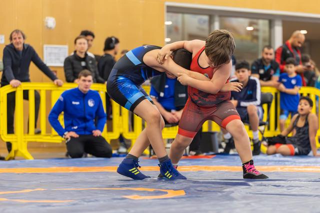 Two young wrestlers locked in a fierce clinch on the mat, leaning into each other with full effort, coaches watching nearby.