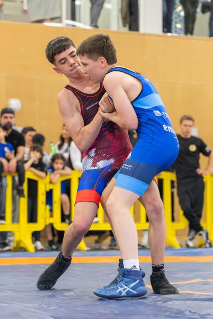 Two teenage wrestlers locked in a tight clinch, both straining with effort on the orange mat before a crowded gymnasium.