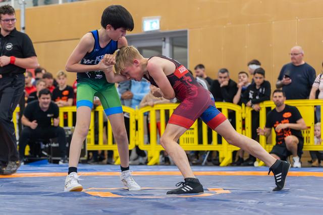 Two young wrestlers face off on a blue mat, one in blue-green, one in red-black, both low and tense.