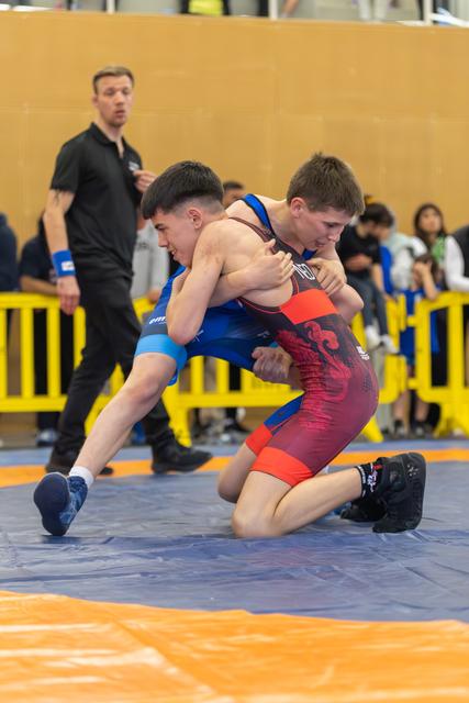 Two young wrestlers locked in a fierce takedown attempt on the mat, referee watching closely in the background.