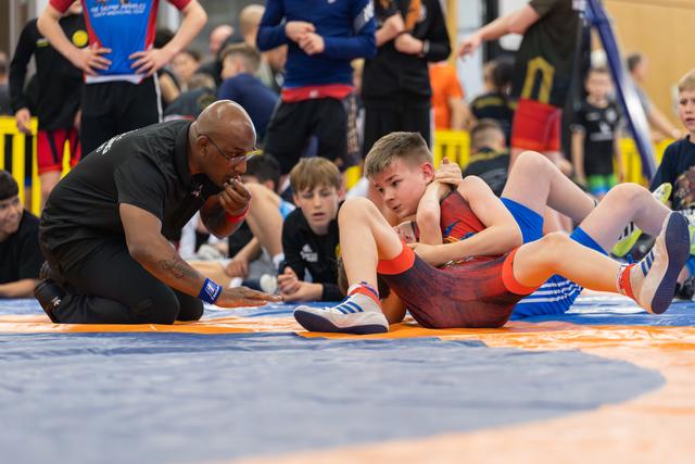 Young wrestler in blue pins opponent on orange mat while coach crouches nearby, watching intently with focused expression.