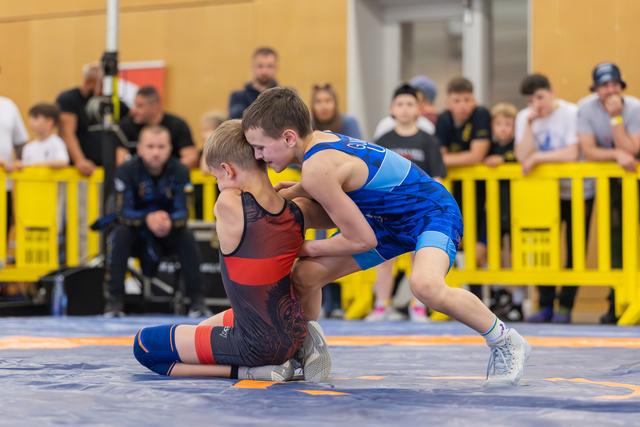 Two young boys wrestling on a blue mat, one in blue singlet dominating over opponent in black, crowd watching.