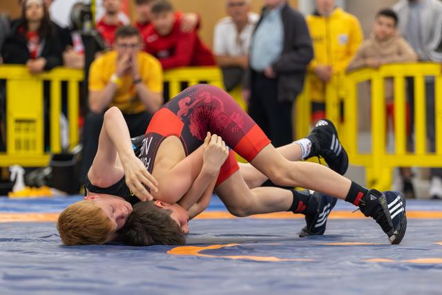 Two young wrestlers locked in a tight hold on the mat, one pinning the other as spectators watch intently behind barriers.